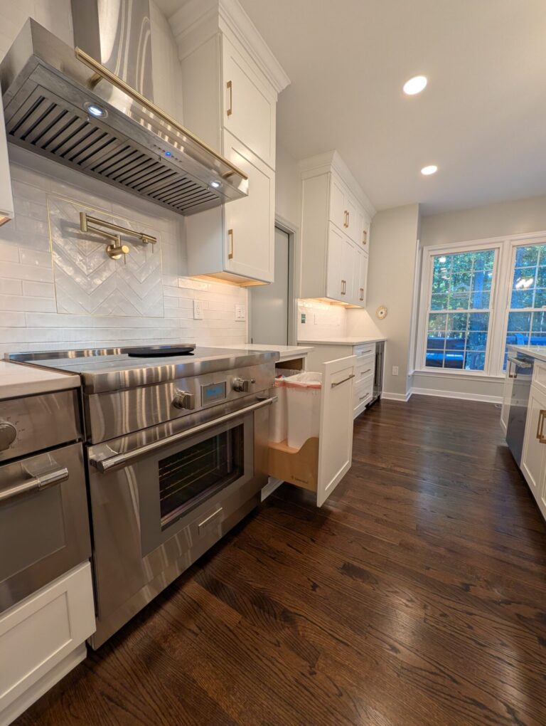 Professional range under a stainless hood with a brass pot filler and herringbone tile inset, beside a pull-out trash cabinet in white shaker cabinetry